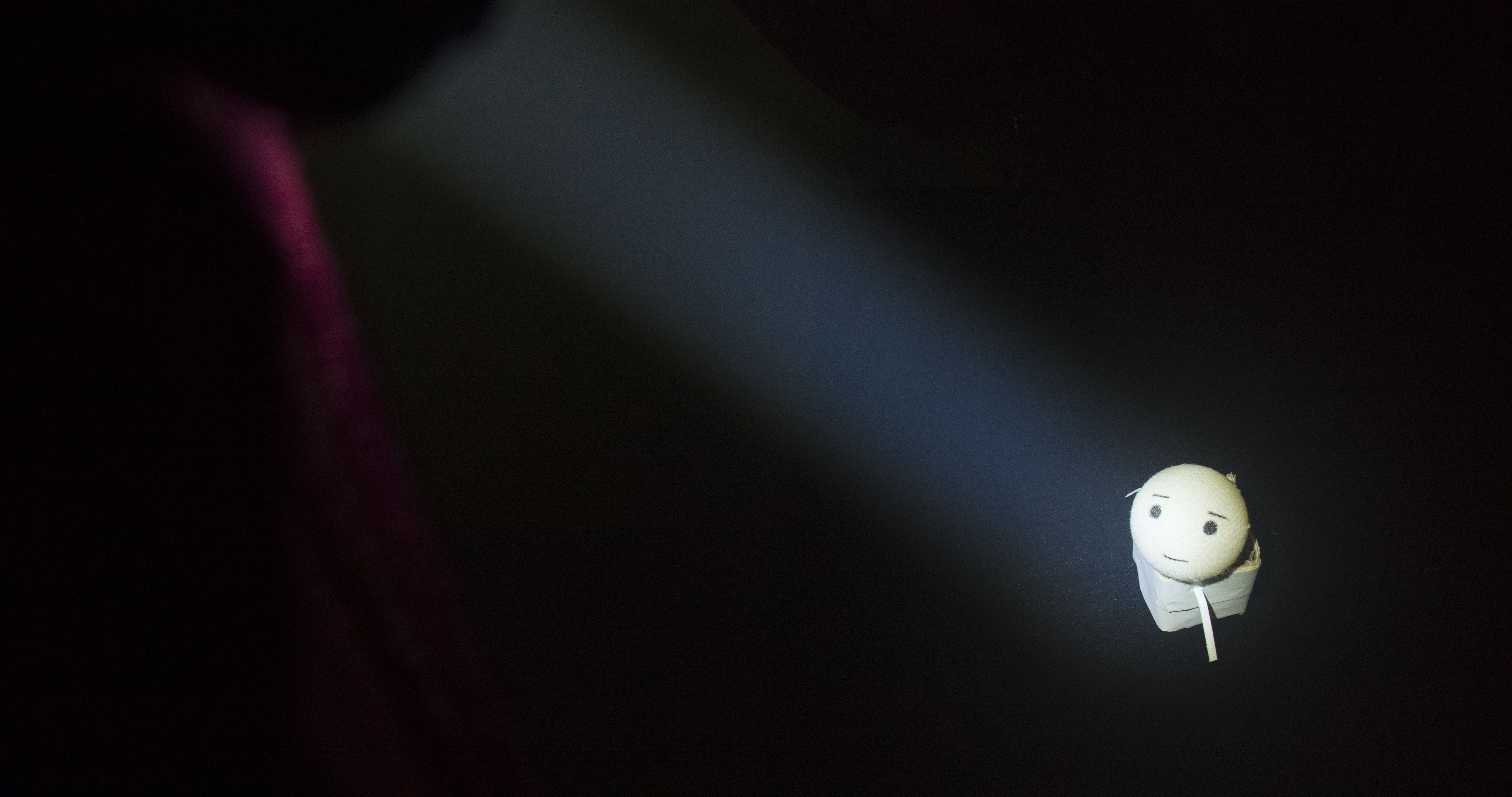 A small handmade figure in a pool of blue stage light, surrounded by darkness